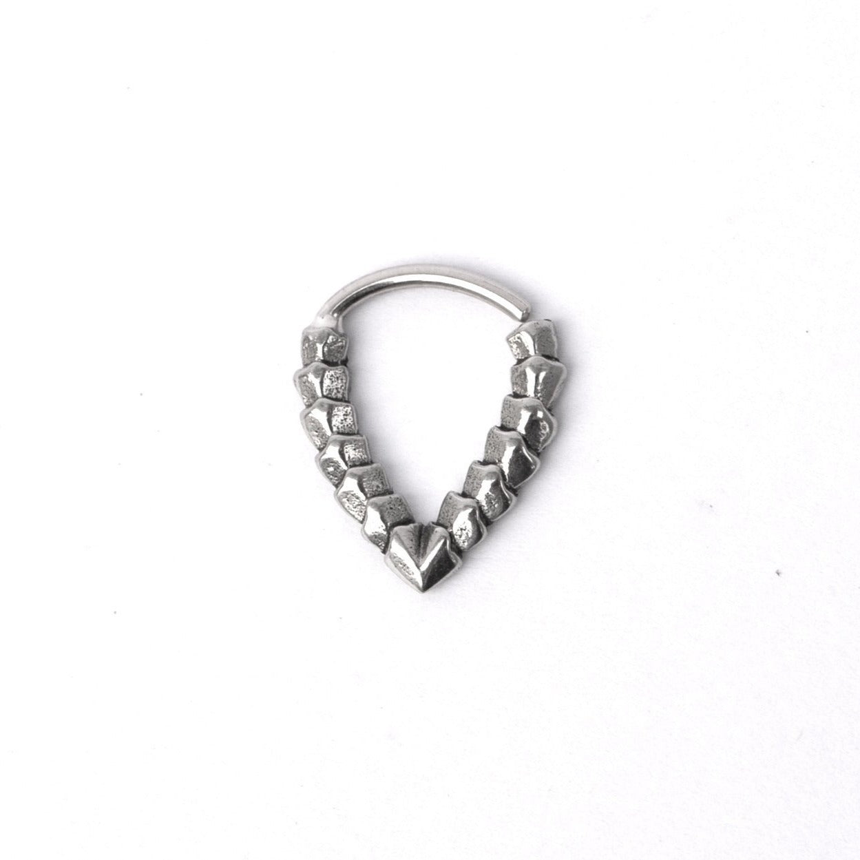 The KAMO Biomech Teardrop Clicker Septum Ring in silver, crafted by Kaya Movement, features a V-shaped design adorned with a series of small, square-like beads along the inner side that create a patterned and textured appearance. This 16-gauge piece of handcrafted body jewelry is displayed against a plain white background.